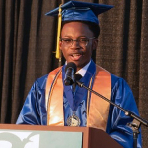 An informal portrait of young graduate in cap and gown, he is at a podium speaking to an audience