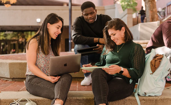 Three students, two women looking at a laptop together, and one man looking over their shoulders. They are in a deep discussion.